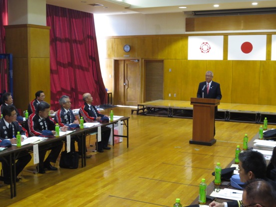 左側の西会津町消防団の皆さんと、右側の消防職員の方々が長机が並んだ席に向かい合って座り、中央の演壇で挨拶を行っている市長の話を聞いている写真