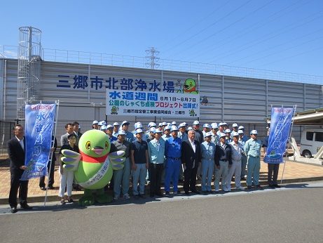 壁に三郷市北部浄水場と書かれた浄水場の前でマスコットキャラクターかいちゃん、市長を中央に並んでいる作業員の方々の集合写真