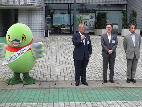 建物入口の前で、マスコットキャラクターかいちゃんの右側に2名の男性が横一列に並び、中央に立っている市長が右手でマイクを持ち挨拶を行っている写真