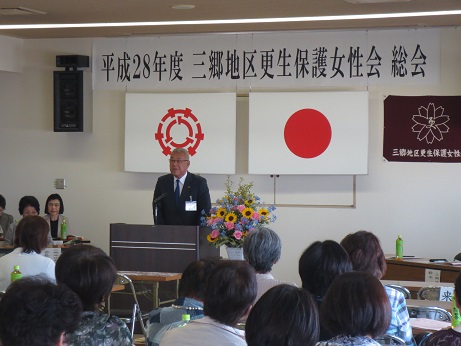 「平成28年度三郷地区更生保護女性会総会」と書かれた横断幕と国旗と三郷市旗が飾られた室内に並んだ長机の席に座っている参加者と、向かい合って演壇に立っている市長が挨拶を行っている写真