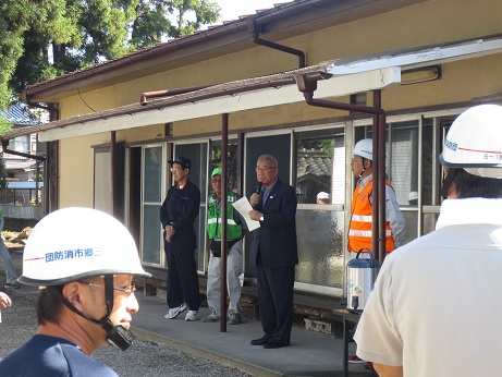 彦川戸公民館前で、防災訓練の挨拶の言葉を述べる三郷市長と話を聞く「三郷市消防団」と書かれている白いヘルメットを着用した関係者の写真