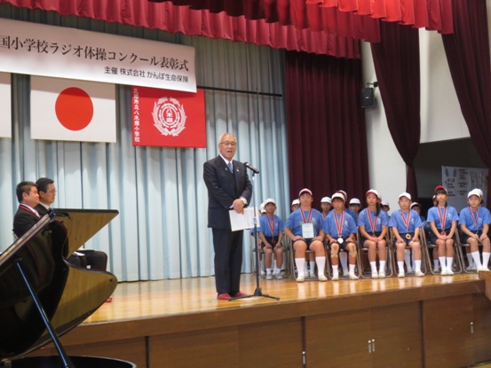 「第4回全国小学校ラジオ体操コンクール表彰式」と書かれた横断幕と国旗・三郷市旗・八木郷小学校の校章旗が吊り下げられているステージの上で、資料を手に持ち話をする三郷市長とパイプ椅子に座り話を聞く紅白帽と体操服を着用し銅メダルを首に掛けた八木小学校の子どもたちの写真