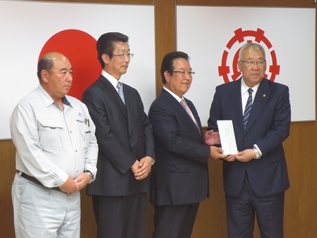 国旗と三郷市旗が飾られた室内で、右側の市長と男性が一緒に頂いた義援金を持ち、左側に2名の関係者の男性が横一列に並んで立っている写真