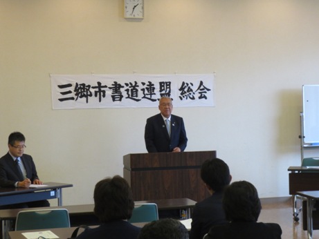 「三郷市書道連盟定期総会」と書かれた横断幕が壁に貼ってあり、その前に設置された演台に立ってお祝いの挨拶を述べる三郷市長と、座って話を聞く書道連盟関係者の写真