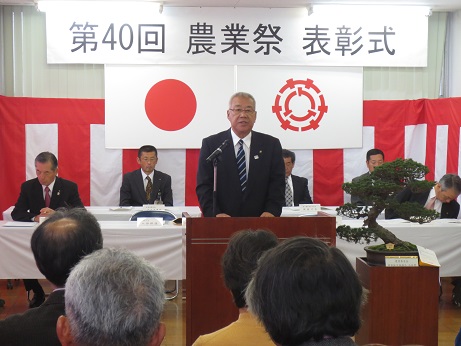 「第40回三郷市農業祭表彰式」と書かれた横断幕と国旗と三郷市旗に紅白幕が貼られた室内で、長机の席に座っている関係者と参加者が向かい合って座り、中央の演壇で挨拶を行っている市長の写真
