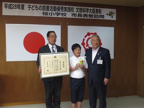 横断幕と国旗と三郷市旗が飾られた室内で、額入りの表彰状を両手で持った小河校長先生と市長の真ん中で、マスコットキャラクターかいちゃんのぬいぐるみを持っている菊地君との記念写真