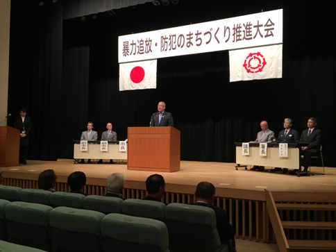 「暴力追放・防犯のまちづくり推進大会」と書かれた横断幕と国旗と三郷市旗が飾られたステージの左右に設置された長机の席に3名ずつ関係者が座り、中央の演壇に立つ市長が挨拶を行っている様子の写真