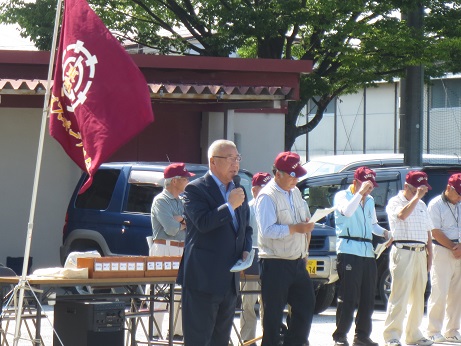 長机にワインレッド色の旗が設置され、右側にお揃いのキャップを被った男性たちが横一列に並んでいる前で、右手にマイクを持ち挨拶を行っている市長の写真