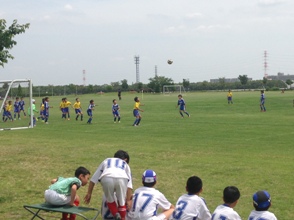 黄色と青色のユニホームを着たチームの子どもたちがサッカーゴールの前で対戦している様子を、手前の椅子に座っている白色のユニホームを着た子供たちが見ている後ろ姿の写真