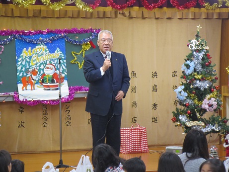 クリスマスツリーなどで飾られた室内でマイクを持ち挨拶を行っている市長の話を座って聞いている子供たちの後ろ姿の写真