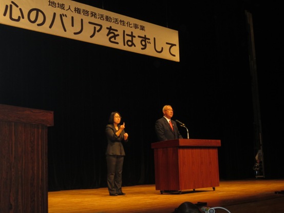 「地域人権啓発活動活性化事業 心のバリアをはずして」と書かれた横断幕下のステージに黒色のスーツを着た手話通訳者の女性が手話を行いながら、演壇で挨拶を行っている市長の写真