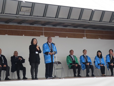 産業フェスタ2017で挨拶の言葉を述べる青い法被を着用した三郷市長と手話通訳をする女性、その後方に並んだパイプ椅子に座り話を聞く青・緑・黒の法被を着用した関係者たちの写真
