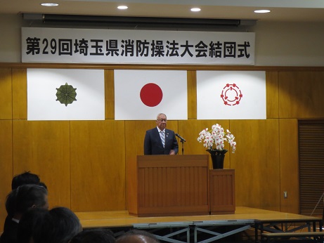「第29回埼玉県消防操法大会結団式」と書かれた横断幕と3枚の旗が飾られた室内の演壇に立ち挨拶を行っている市長の写真