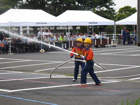 黄色のヘルメットにオレンジ色と紺色の作業着を着た2名の消防クラブ員の子どもがホースを握り、消防ポンプ操法を行っている様子の写真