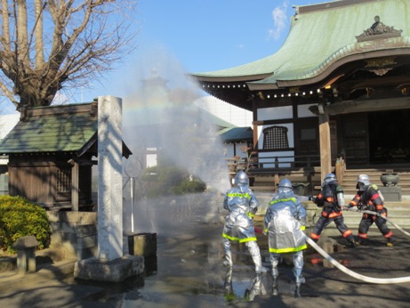玉蔵院の建物と左側の祠の間に向かって消防団員が放水を行っている訓練の様子の写真