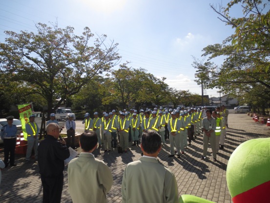 駐車場横の歩道で、6列に整列して立っている薄いグリーンの作業着に安全反射ベストと帽子を着用した、たくさんの参加者に向かって、マイクを手に持ち話をする三郷市長を背後から写した写真