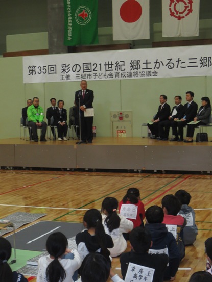 「第35回彩の国21世紀郷土かるた三郷」と書かれたボードが設置されている体育館で、座っている子どもたちが舞台上に立って挨拶をしている市長の方を見ている写真