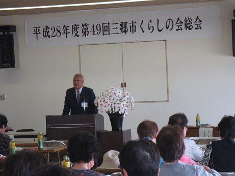 「平成28年度第49回三郷市くらしの会総会」と書かれた横幕が壁に貼られ、室内に設置された長机の席に座っている参加者と向かい合って演壇に立っている市長が挨拶を行っている写真