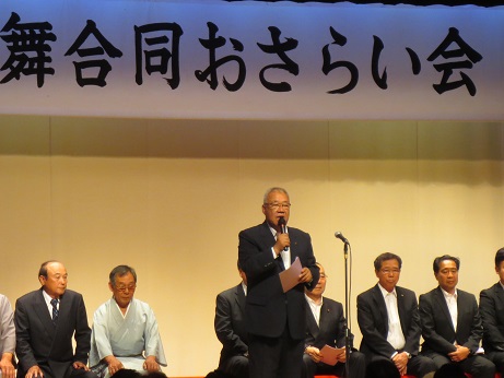 「第37回民謡・民舞合同おさらい会」と書かれた横断幕の下、関係者が横一列に椅子に座っている前で、右手にマイクを持った市長が挨拶を行っている写真