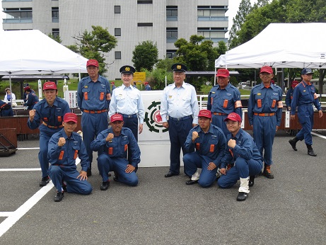 演壇の前で、市長と左側の男性を挟んで左右に各4名ずつ消防団の男性が並んでいる記念写真