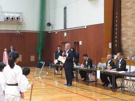 ステージ下に長机が並んで設置されている体育館で、右手にマイクを持って挨拶を行っている市長の話を、道着を着用した子供たちが聞いている後ろ姿の写真