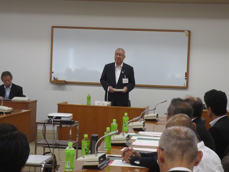 ホワイトボードの前に配置された演台に立ちあいさつの言葉を述べる三郷市長と、ペットボトルのお茶とマイクが置かれた席に座り話を聞く参加者を、参加者後方から撮影した写真