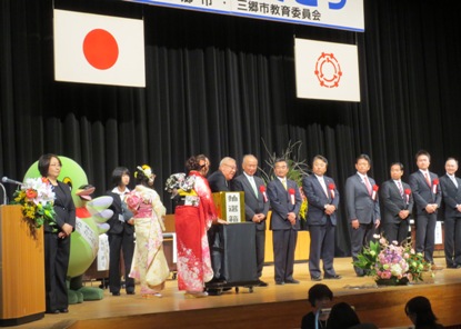 胸章リボンをつけた関係者の方々が横一列に並んだステージで、金色の紙が貼られた抽選箱の中に手を入れ抽選を行っている市長の写真