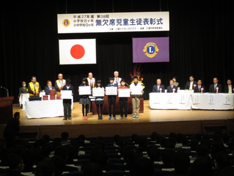 「第38回無欠席児童生徒表彰式」と書かれた横断幕下のステージ上の両端の席に市長をはじめ胸章リボンをつけた関係者が座り、演壇の前に6名の子どもたちが賞状を持って並んでいる写真