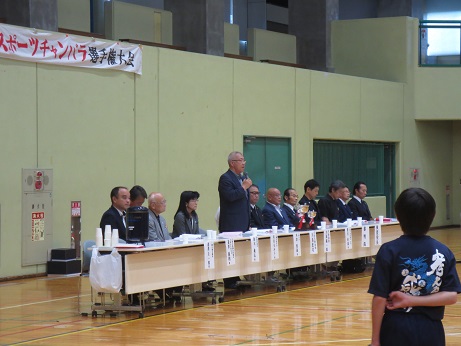 「第10回三郷市スポーツチャンバラ選手権大会」と書かれた横断幕が掛かった壁を背に、中央に2つのトロフィーが置かれた長テーブルの席に立って、マイクを手に持ちあいさつの言葉を述べる三郷市長と、各席に座り話を聞く関係者の写真