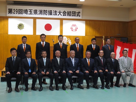 「第29回埼玉県消防操法大会結団式」と書かれた横断幕と3枚の旗が飾られた室内で、前列に設置された椅子に市長を中央に左に4名右に4名の男性が座り、2列目に6名の男性が立っている記念写真