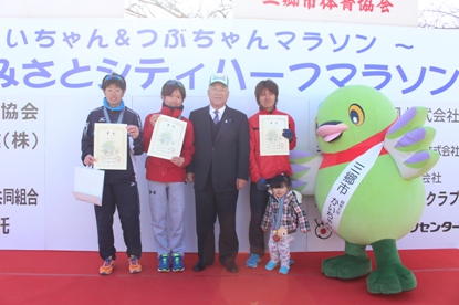 ステージ上で市長を挟んで賞状を手に持った3名のランナーと女の子、マスコットキャラクターかいちゃんが並んで立っている表彰式の様子の写真