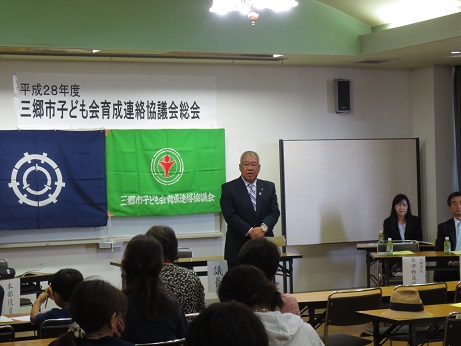 「平成28年度三郷市子ども会育成連絡協議会総会」と書かれた横幕と2枚の旗が飾られた室内に並んだ長机の席に座っている参加者と向かい合って立っている市長が挨拶を行っている写真