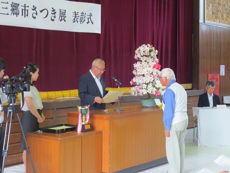 水色のシャツを着た男性と市長が演壇を挟んで向かい合って立ち、表彰状の授与を行っている様子を左からテレビカメラで撮影している様子の写真