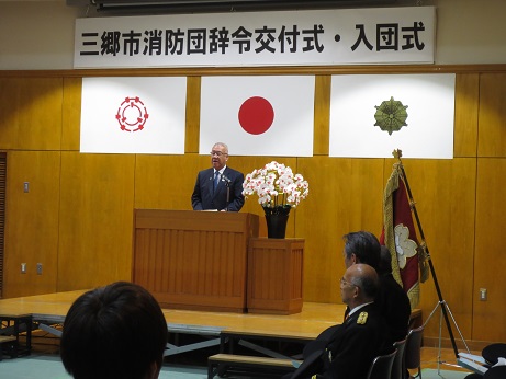 三郷市旗・国旗・消防旗の上に「三郷市消防団辞令交付式・入団式」と書かれた横断幕が吊り下げられ、白い花が飾られた花台と並ぶ演台に立ちあいさつの言葉を述べる三郷市長の写真