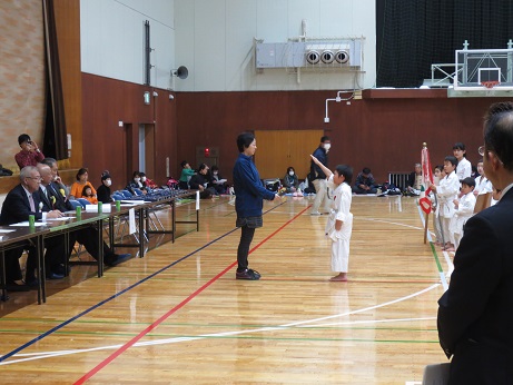 選手たちの前で女性と道着を着用した男の子が向かい合って立ち、男の子が右手を真っ直ぐ上にあげて選手宣誓を行っている様子の写真