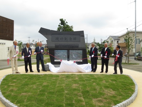 竣功記念碑と書かれた記念碑の手前で左右に赤色の紐がつけられた白布を左右に各3名の関係者が引っ張り除幕が行われた直後の様子の写真