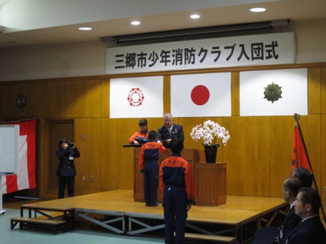 三郷市旗・国旗・消防旗の上に「三郷市少年消防クラブ入団式」と書かれた横断幕が吊り下げられ、白い花が飾られた花台と並ぶ演台に立ち制服を着用した入団者に少年消防手帳とクラブ員章を交付している三郷市長の写真