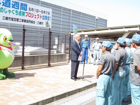 「水道週間6月1日～6月7日公園のじゃぐち点検プロジェクト出陣式！」と書かれた横断幕が固定されているフェンスの前で、挨拶の言葉を述べる三郷市長と向かい合って立ち話を聞く三郷市指定管工事業協同組合のメンバー、タスキをかけたかいちゃんの写真