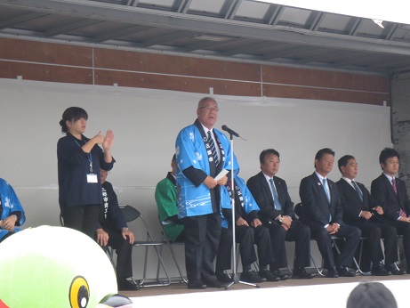 小さなステージの後方に設置された椅子に関係者が座り、手話通訳者の女性の隣でマイクスタンドの前に立っている市長が挨拶を行っている写真