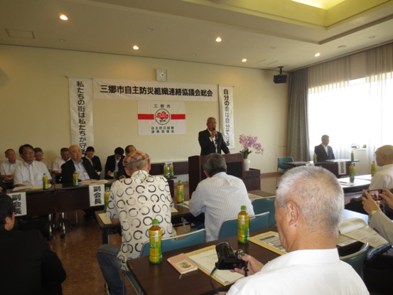 「三郷市自主防災組織連絡協議会総会」と書かれた横断幕が壁に貼り付けてあり、ピンク色の花々で彩られた花台と並ぶ演台に立ってあいさつの言葉を述べる三郷市長と、ペットボトルのお茶と資料が置かれた席に座り話を聞く参加者の写真