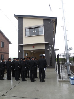 2階建ての機械器具置場の建物の方を見て並んで立っている消防団員の皆さんの後ろ姿の写真
