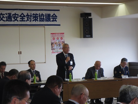 「三郷市交通安全対策協議会」と書かれた横幕が貼られた室内で、右手にマイクを持ち挨拶を行っている市長の写真