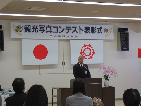 「観光写真コンテスト表彰式」と書かれた横断幕と国旗と三郷市旗が飾られた会場の檀上で挨拶を行っている市長の写真