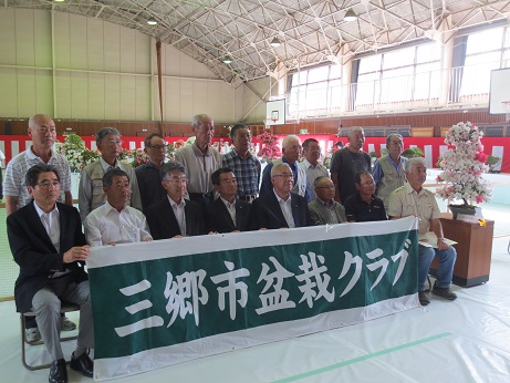 「三郷市盆栽クラブ」と書かれた横断幕の後方の前列に市長を中央に左に4名右に3名の男性が椅子に座り、二列目に9名の男性が並んで立っている記念写真