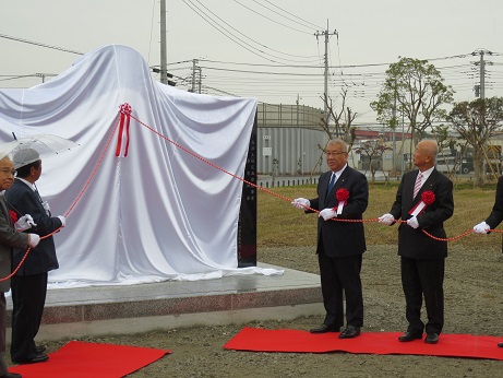 竣功記念碑に掛けられた白幕に取り付けられた赤色の紐を胸章リボンをつけた市長や関係者が握っている除幕式の様子の写真