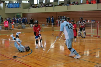 水色のユニホームを着た選手がパックのそばで頭と背中を床に打ちつけて転び、近くで赤色と水色のユニホームを着たチームが対戦している写真