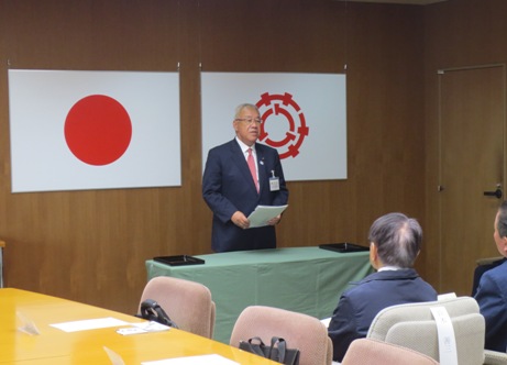 国旗と三郷市旗が飾られた室内で、椅子に座っている委嘱状を交付された方々と向かい合った市長が挨拶を行っている写真
