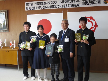 横断幕と国旗と三郷市旗が飾られた室内で、市長を挟んで1名の男児と2名の男子生徒、1名の女子生徒が笑顔で盾と透明袋に入った小さなかいちゃんのぬいぐるみを持っている記念写真