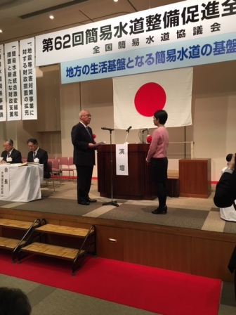 「第62回簡易水道整備促進全国大会」と書かれたボードの下に「地方の生活基盤となる簡易水道の基盤強化」と書かれたボードと国旗が吊るしてあるステージの演台前で、受賞者を代表し向かいあって立つ女性へ謝辞を述べる三郷市長の写真