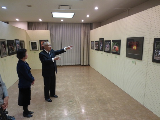 両側に配置された白い展示パネルに作品が横一列に飾ってあり、三郷市長が夕日を映した写真を指差しながら隣に立つ女性に感想を述べている写真